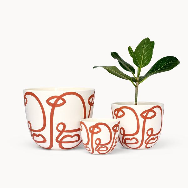 Three hand-painted vessels in white with free-flowing curves in terracotta painted on them, resembling faces. A plant is standing in one of the vessels.