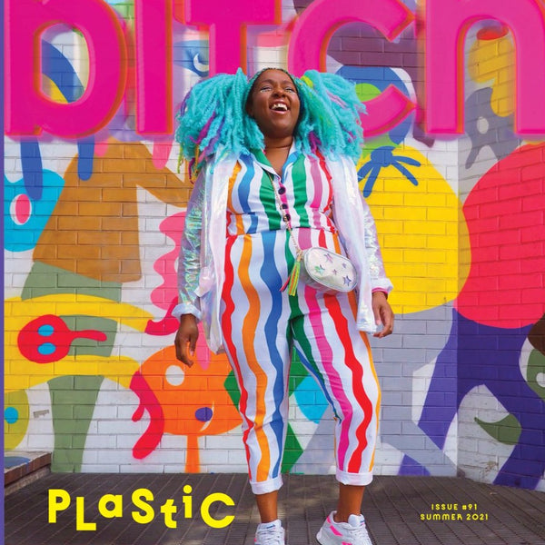 A black woman with mint hair is laughing in front of a wall of color street art. She is wearing a bright, vertically striped suit.