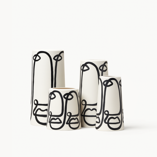 Three tall hand-painted vessels in white with free-flowing curves in black painted on them, resembling faces. A plant is standing in one of the vessels.